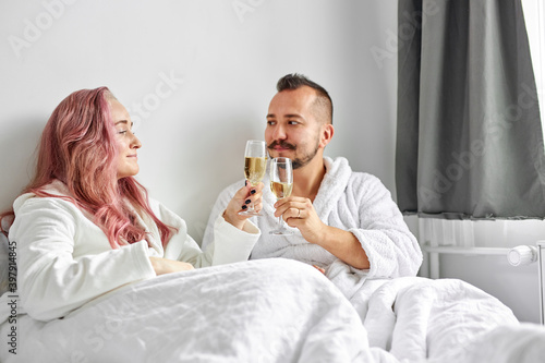happy young caucasian couple relaxing in bed, drinking sparkling champagne from glasses. celebrating. weekend, rest, relax