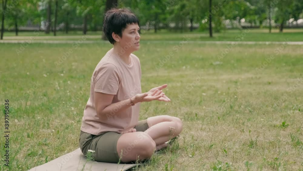 Wide shot of sporty middle-aged Caucasian woman with dark short hair wearing sportswear sitting on yoga mat on grass outdoors and speaking Wide shot of sporty middle-aged Caucasian woman with dark short hair wearing sportswear sitting on yoga mat on grass outdoors and speaking