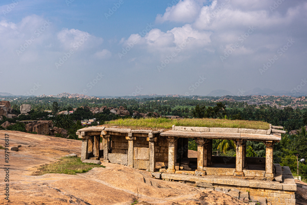 Hampi, Karnataka, India - November 4, 2013: Virupaksha Temple complex. Moola brown stone gallery ruin with grass growing on top under light blue cloudscape and green foliage around.