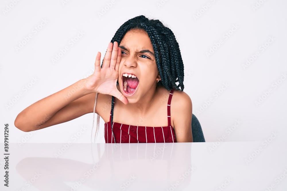 Young african american girl child with braids wearing casual clothes sitting on the table shouting and screaming loud to side with hand on mouth. communication concept. Young african american girl child with braids wearing casual clothes sitting on the table shouting and screaming loud to side with hand on mouth. communication concept.