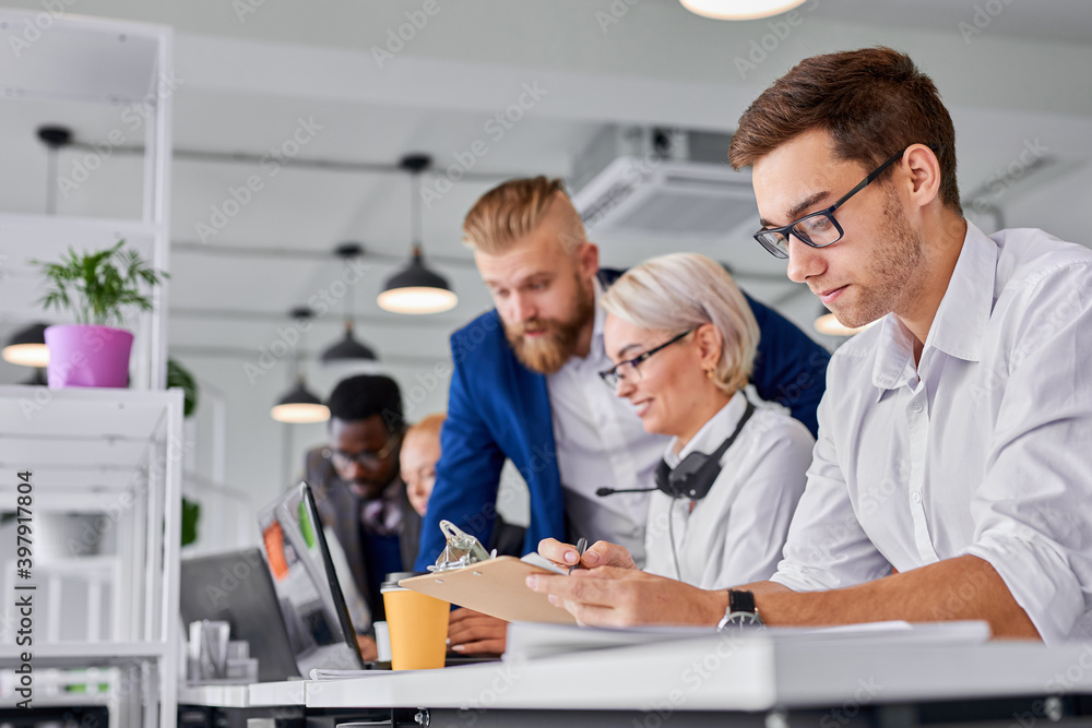 manager male is working in the office, caucasian guy looking at paper concentrated, thinking, while others are coworking together, focus on man