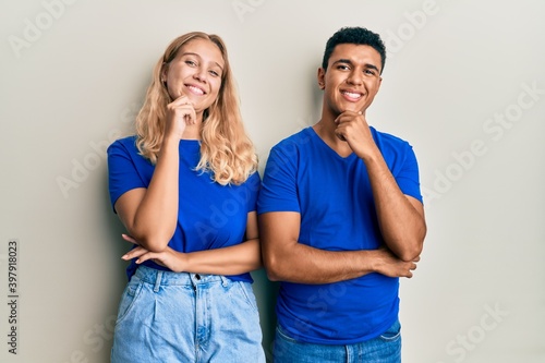 Young interracial couple wearing casual clothes looking confident at the camera smiling with crossed arms and hand raised on chin. thinking positive.