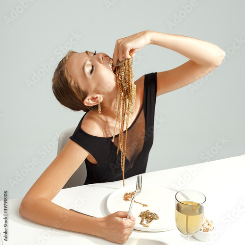 A young attractive stylish girl eats gold jewelry from a plate. creative idea of consumerism