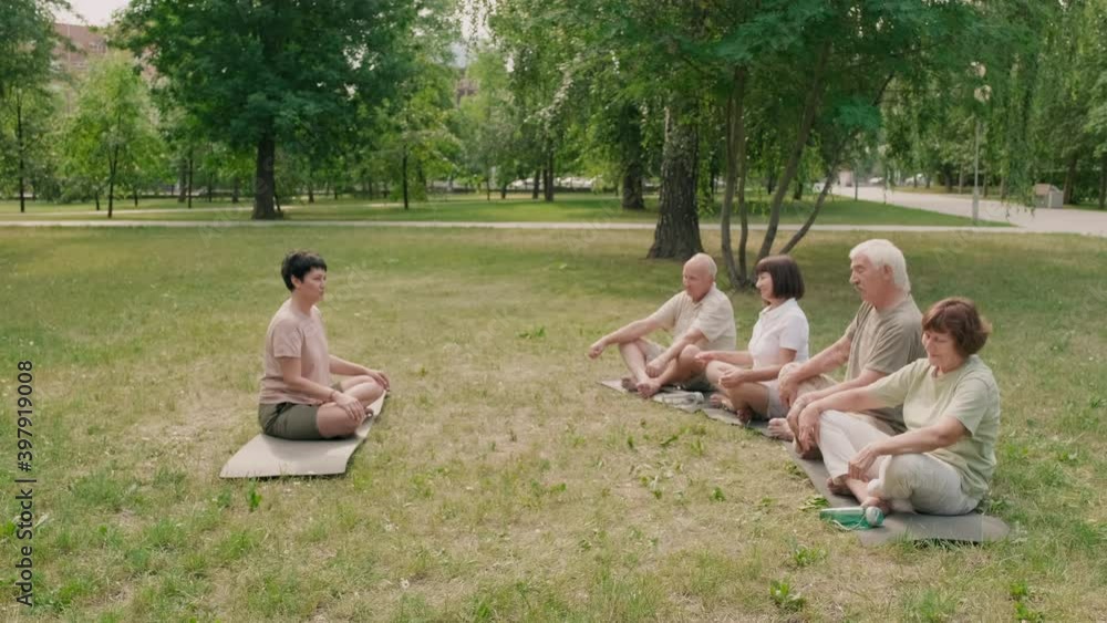Wide shot of middle-aged Caucasian female yoga coach sitting in lotus pose on mat on grass in park and talking to four senior people sitting in front of her Wide shot of middle-aged Caucasian female yoga coach sitting in lotus pose on mat on grass in park and talking to four senior people sitting in front of her