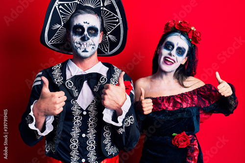 Young couple wearing mexican day of the dead costume over red success sign doing positive gesture with hand, thumbs up smiling and happy. cheerful expression and winner gesture.