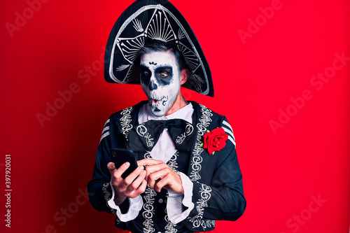Young man wearing mexican day of the dead costume using smartphone scared and amazed with open mouth for surprise, disbelief face