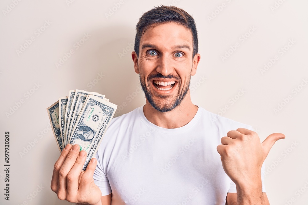 Young handsome man holding bunch of dollars banknotes over isolated white background pointing thumb up to the side smiling happy with open mouth Young handsome man holding bunch of dollars banknotes over isolated white background pointing thumb up to the side smiling happy with open mouth