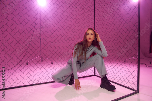 Fashion model in green sporty clothes and black boots sitting on white florr against grid fence and pink background. Girl with long straight brunette hair photo