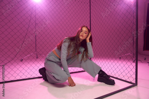 Fashion model in green sporty clothes and black boots sitting on white florr against grid fence and pink background. Girl with long straight brunette hair photo