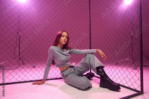 Fashion model in green sporty clothes and black boots sitting on white florr against grid fence and pink background. Girl with long straight brunette hair photo