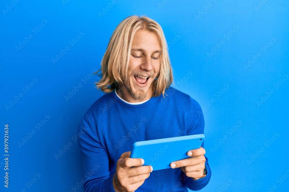 Caucasian young man with long hair using touchpad device smiling and laughing hard out loud because funny crazy joke. Caucasian young man with long hair using touchpad device smiling and laughing hard out loud because funny crazy joke.