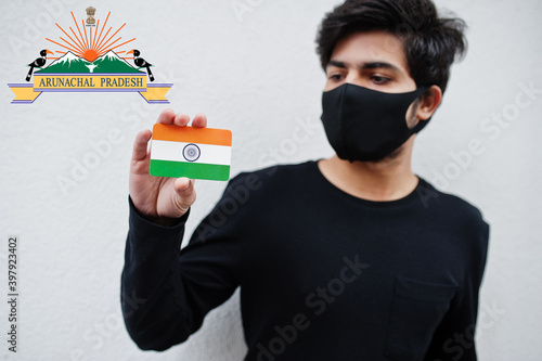 Indian man wear all black and face mask, hold India flag in hand isolated on white background with Arunachal Pradesh state emblem . Coronavirus India states and union territories concept. photo