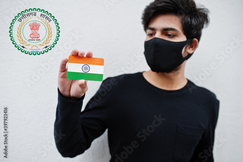 Indian man wear all black and face mask, hold India flag in hand isolated on white background with Chhattisgarh state emblem . Coronavirus India states and union territories concept. photo