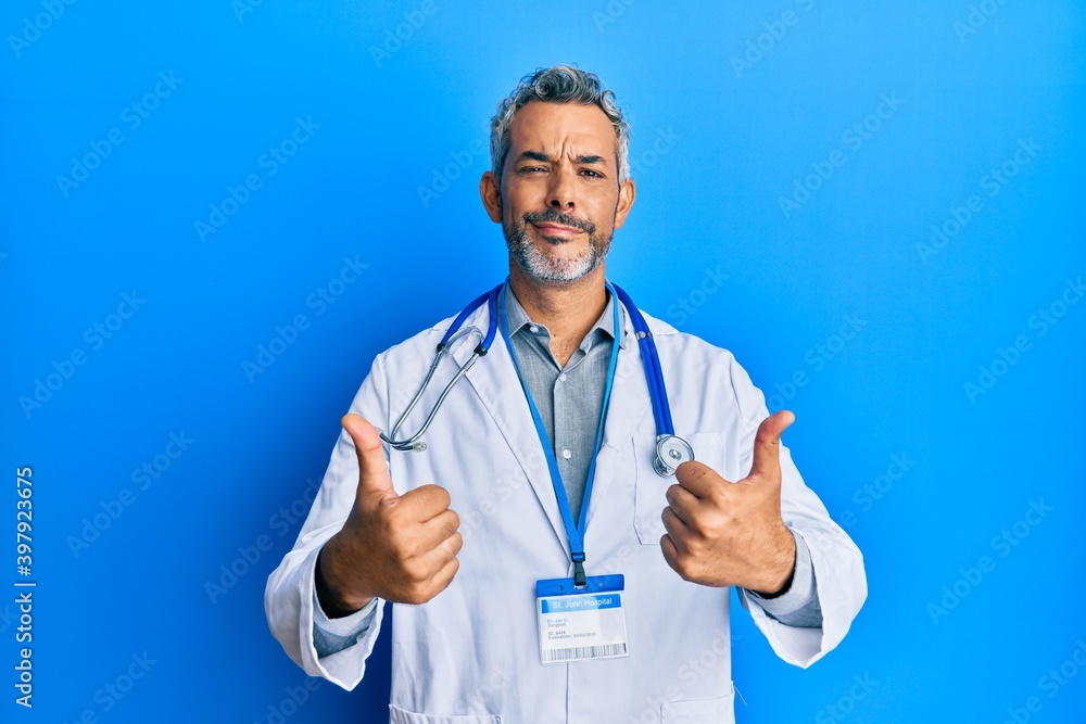 Middle age grey-haired man wearing doctor uniform and stethoscope approving doing positive gesture with hand, thumbs up smiling and happy for success. winner gesture. Middle age grey-haired man wearing doctor uniform and stethoscope approving doing positive gesture with hand, thumbs up smiling and happy for success. winner gesture.