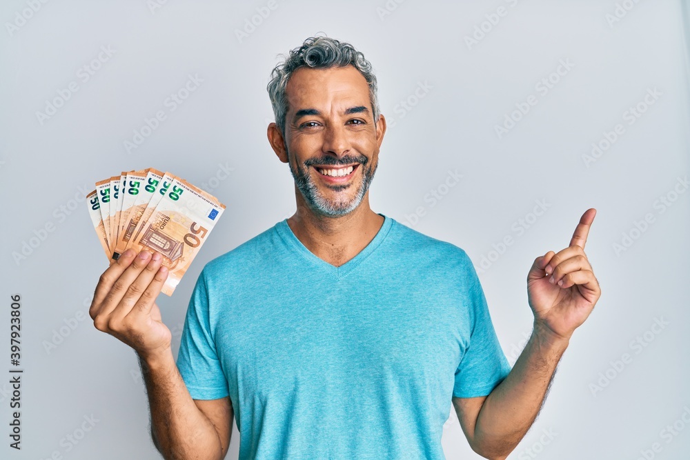 Middle age grey-haired man holding 50 euro banknotes smiling happy pointing with hand and finger to the side Middle age grey-haired man holding 50 euro banknotes smiling happy pointing with hand and finger to the side