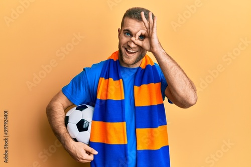 Young caucasian man football hooligan cheering game holding ball smiling happy doing ok sign with hand on eye looking through fingers