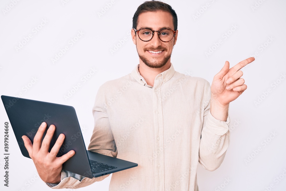 Young handsome man holding laptop smiling happy pointing with hand and finger to the side Young handsome man holding laptop smiling happy pointing with hand and finger to the side