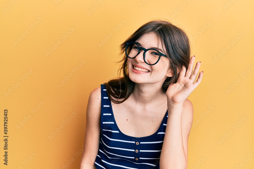 Young beautiful caucasian girl wearing casual clothes and glasses smiling with hand over ear listening and hearing to rumor or gossip. deafness concept.