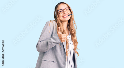 Young beautiful blonde woman wearing call center agent headset doing happy thumbs up gesture with hand. approving expression looking at the camera showing success. photo