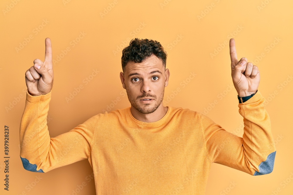 Young hispanic man pointing up with fingers in shock face, looking skeptical and sarcastic, surprised with open mouth Young hispanic man pointing up with fingers in shock face, looking skeptical and sarcastic, surprised with open mouth