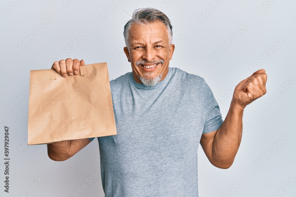 Middle age grey-haired man holding take away paper bag screaming proud, celebrating victory and success very excited with raised arm Middle age grey-haired man holding take away paper bag screaming proud, celebrating victory and success very excited with raised arm