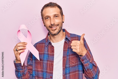 Young handsome man holding pink cancer ribbon smiling happy and positive, thumb up doing excellent and approval sign