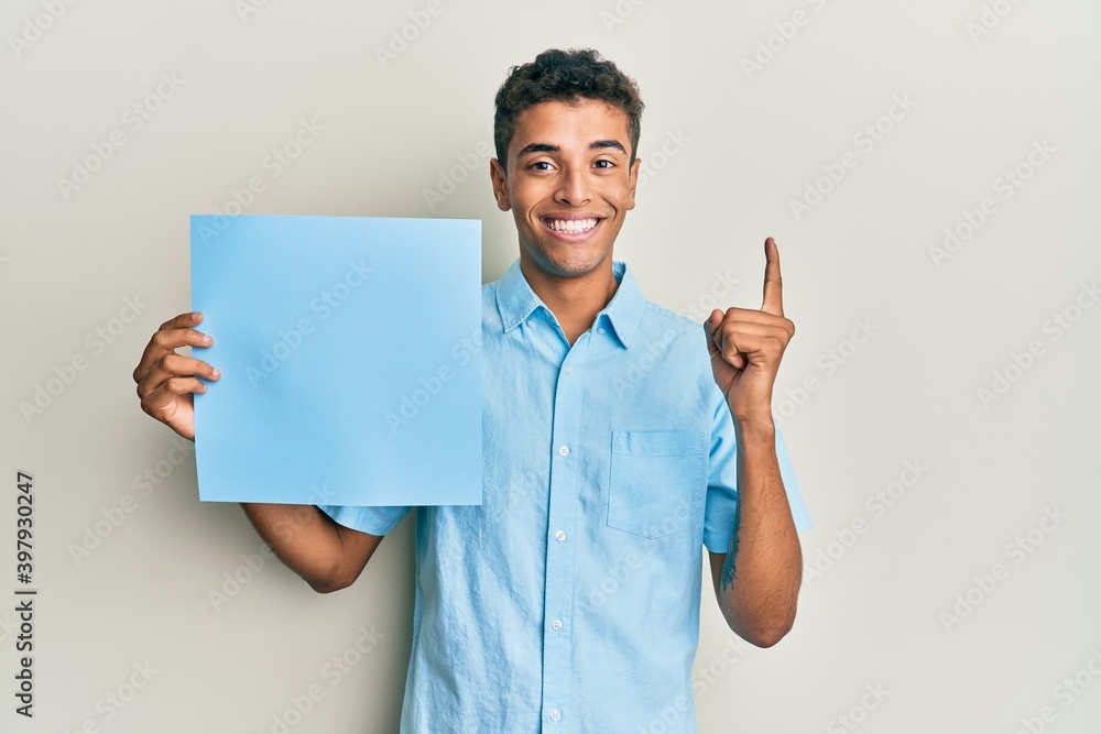Young handsome african american man holding blue blank empty banner smiling with an idea or question pointing finger with happy face, number one Young handsome african american man holding blue blank empty banner smiling with an idea or question pointing finger with happy face, number one