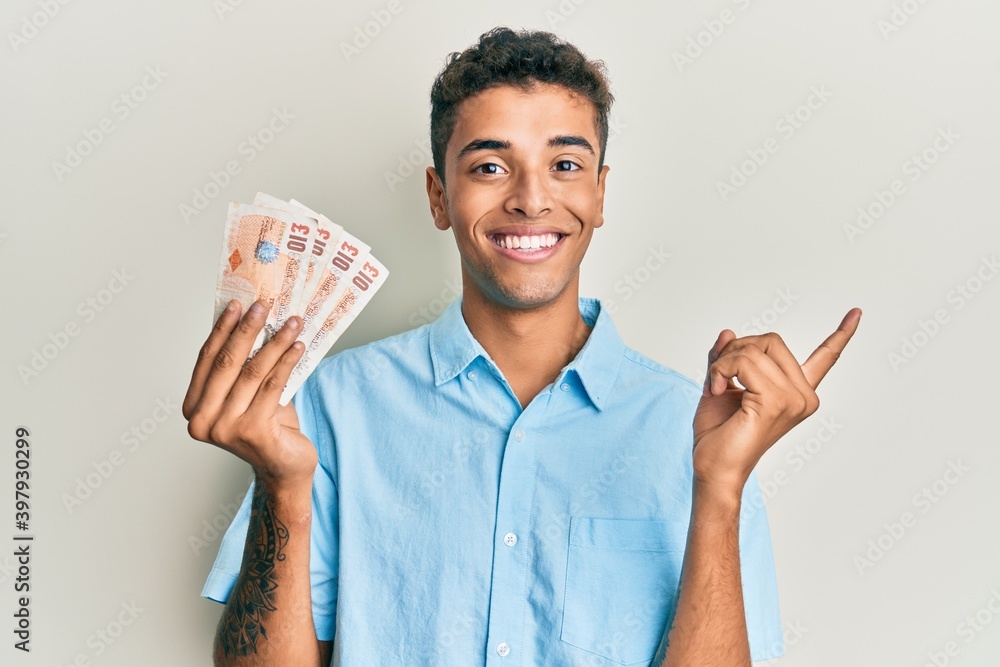 Young handsome african american man holding 10 united kingdom pounds banknotes smiling happy pointing with hand and finger to the side Young handsome african american man holding 10 united kingdom pounds banknotes smiling happy pointing with hand and finger to the side