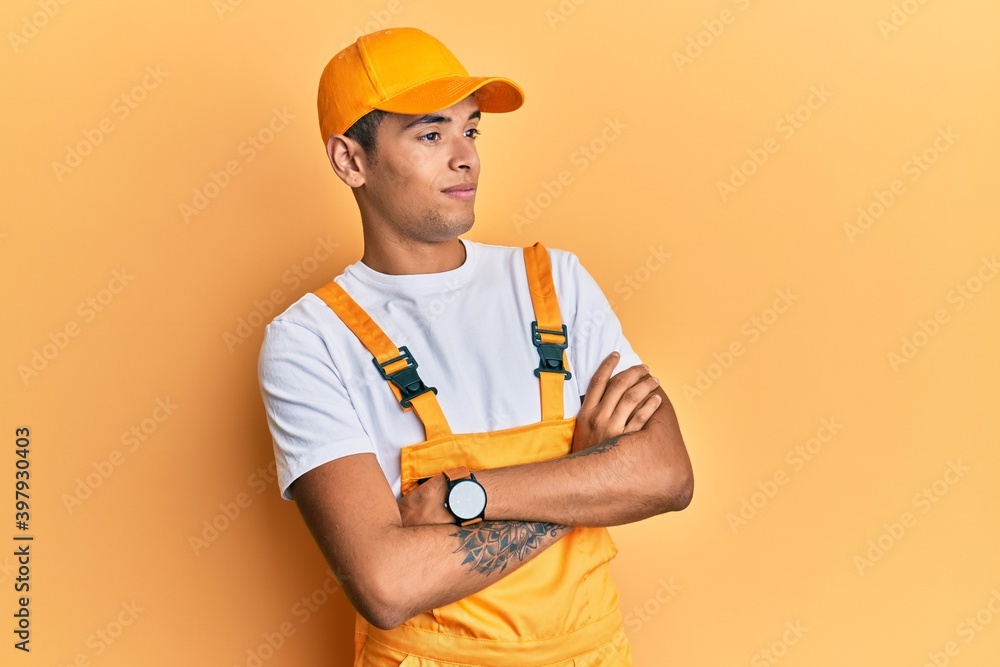 Young handsome african american man wearing handyman uniform over yellow background looking to the side with arms crossed convinced and confident