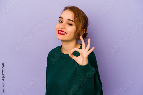 Young arab mixed race woman winks an eye and holds an okay gesture with hand.