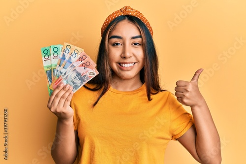 Young latin woman holding australian dollars banknotes smiling happy and positive, thumb up doing excellent and approval sign
