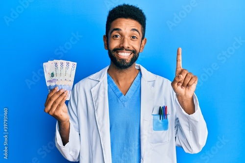 Handsome hispanic man with beard wearing doctor uniform holding 50 colombian pesos smiling with an idea or question pointing finger with happy face, number one © Krakenimages.com