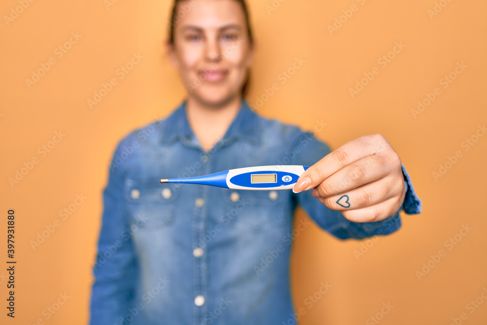 Young beautiful woman smiling happy and confident. Standing with smile on face holding thermometer over isolated yellow background Young beautiful woman smiling happy and confident. Standing with smile on face holding thermometer over isolated yellow background