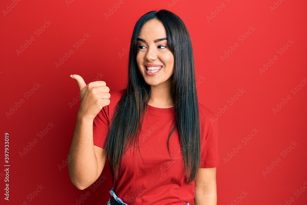 Young beautiful hispanic girl wearing casual red tshirt pointing thumb up to the side smiling happy with open mouth Young beautiful hispanic girl wearing casual red tshirt pointing thumb up to the side smiling happy with open mouth