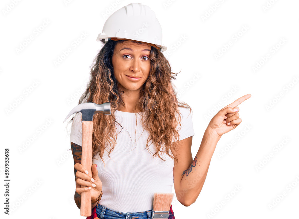 Young hispanic woman with tattoo wearing hardhat and builder clothes holding hammer smiling happy pointing with hand and finger to the side Young hispanic woman with tattoo wearing hardhat and builder clothes holding hammer smiling happy pointing with hand and finger to the side