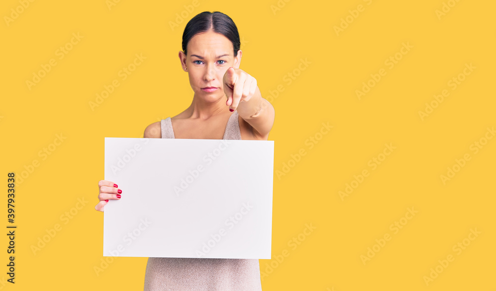 Beautiful brunette young woman holding blank empty banner pointing with finger to the camera and to you, confident gesture looking serious Beautiful brunette young woman holding blank empty banner pointing with finger to the camera and to you, confident gesture looking serious