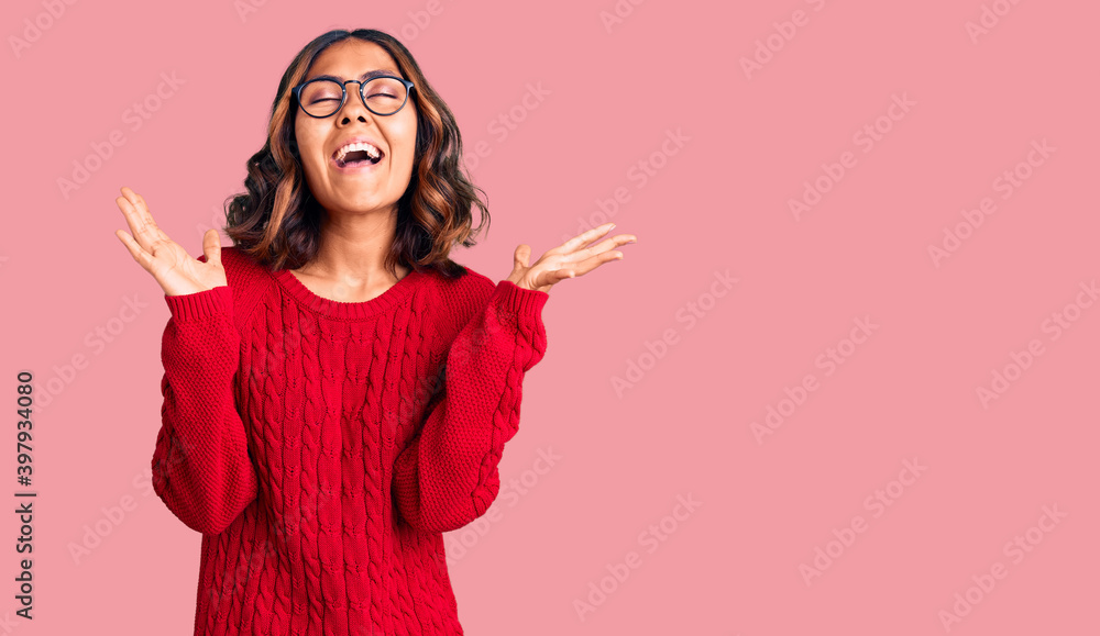 Young beautiful mixed race woman wearing red sweater and glasses celebrating mad and crazy for success with arms raised and closed eyes screaming excited. winner concept Young beautiful mixed race woman wearing red sweater and glasses celebrating mad and crazy for success with arms raised and closed eyes screaming excited. winner concept