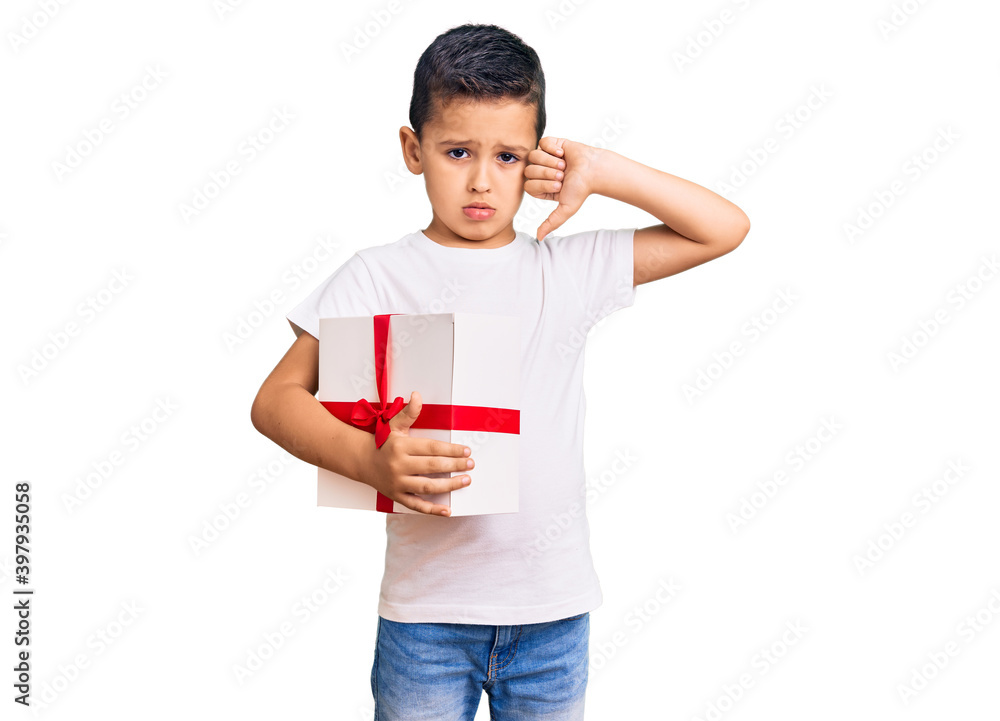 Little cute boy kid holding gift with angry face, negative sign showing dislike with thumbs down, rejection concept Little cute boy kid holding gift with angry face, negative sign showing dislike with thumbs down, rejection concept