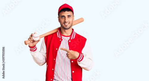Young hispanic man playing baseball holding bat and ball smiling happy pointing with hand and finger