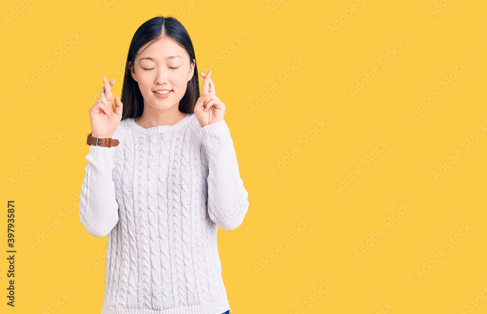 Young beautiful chinese woman wearing casual sweater gesturing finger crossed smiling with hope and eyes closed. luck and superstitious concept. Young beautiful chinese woman wearing casual sweater gesturing finger crossed smiling with hope and eyes closed. luck and superstitious concept.