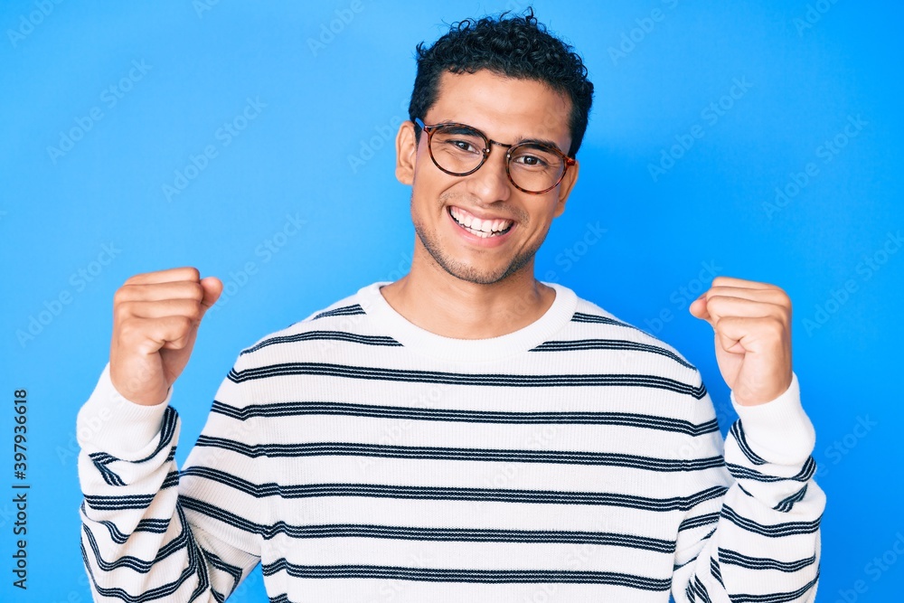 Young handsome hispanic man wearing casual sweater and glasses screaming proud, celebrating victory and success very excited with raised arms Young handsome hispanic man wearing casual sweater and glasses screaming proud, celebrating victory and success very excited with raised arms