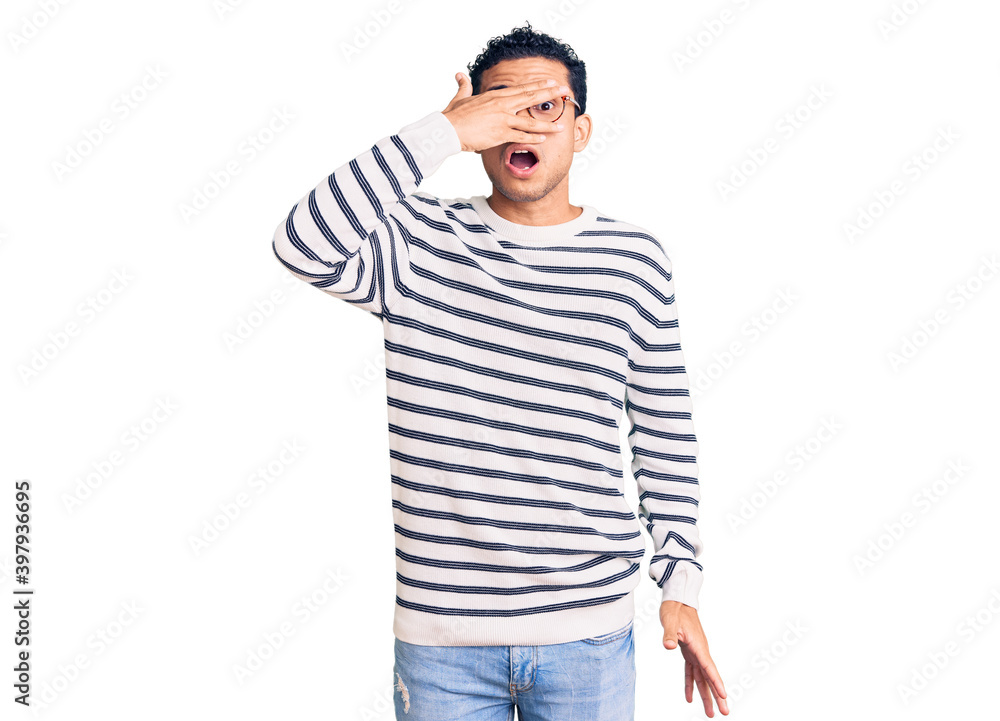 Fototapeta premium Hispanic handsome young man wearing casual clothes and glasses peeking in shock covering face and eyes with hand, looking through fingers with embarrassed expression.