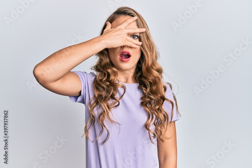 Young blonde girl wearing casual clothes peeking in shock covering face and eyes with hand, looking through fingers afraid