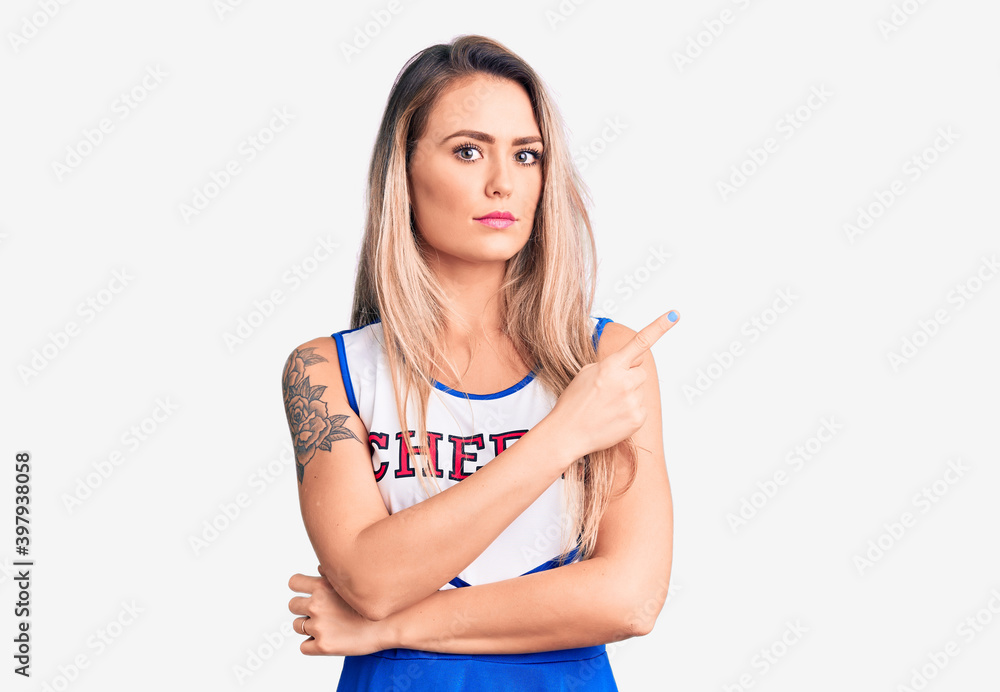 Young beautiful blonde woman wearing cheerleader uniform pointing with hand finger to the side showing advertisement, serious and calm face Young beautiful blonde woman wearing cheerleader uniform pointing with hand finger to the side showing advertisement, serious and calm face