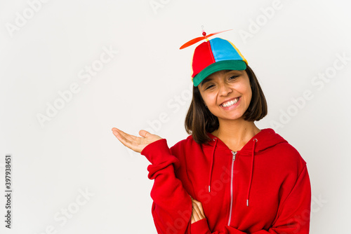 Young hispanic woman wearing a cap with propeller isolated showing a copy space on a palm and holding another hand on waist.
