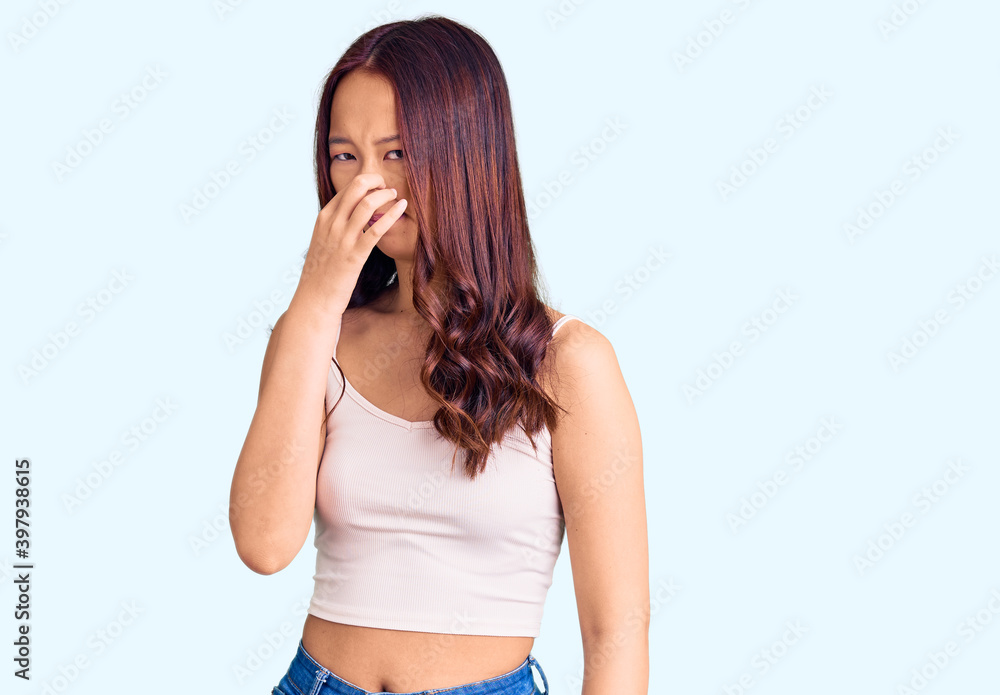 Young beautiful chinese girl wearing casual clothes smelling something stinky and disgusting, intolerable smell, holding breath with fingers on nose. bad smell Young beautiful chinese girl wearing casual clothes smelling something stinky and disgusting, intolerable smell, holding breath with fingers on nose. bad smell