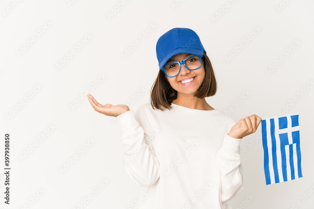 Young hispanic mixed race woman holding a greece flag showing a copy space on a palm and holding another hand on waist. Young hispanic mixed race woman holding a greece flag showing a copy space on a palm and holding another hand on waist.
