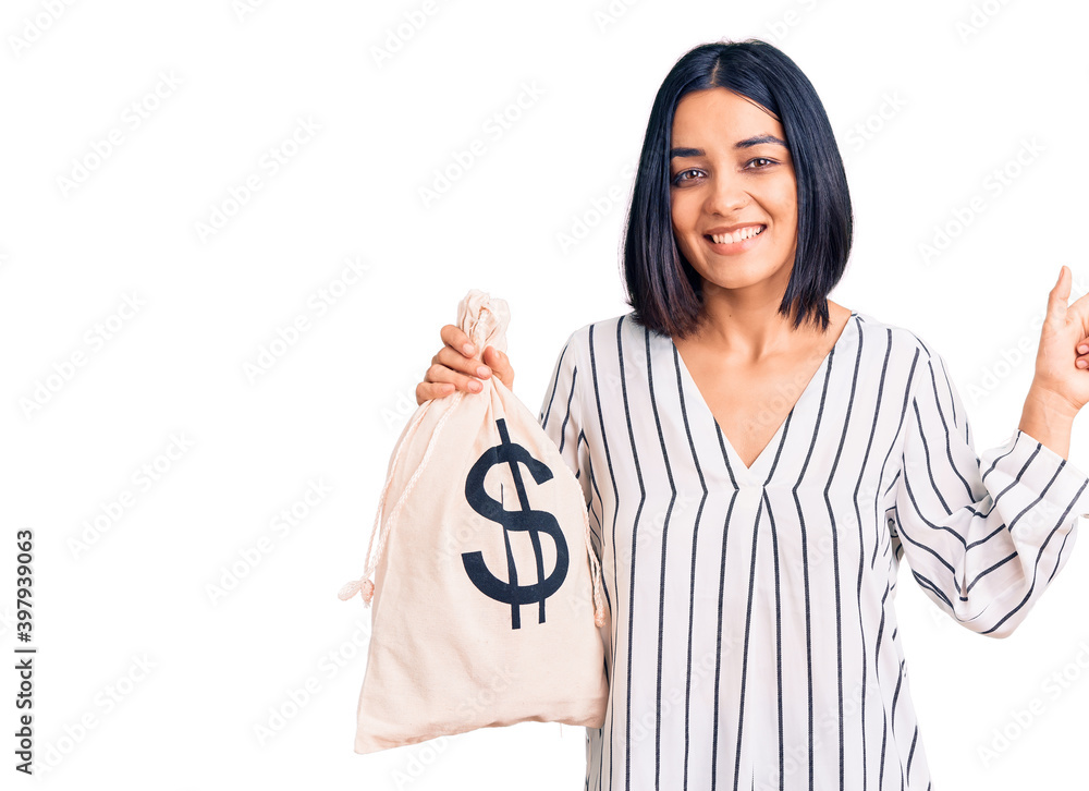 Young beautiful latin woman holding money bag with dollar symbol smiling happy pointing with hand and finger to the side Young beautiful latin woman holding money bag with dollar symbol smiling happy pointing with hand and finger to the side