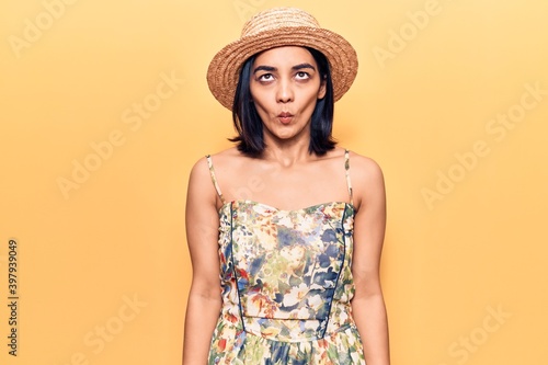 Young beautiful latin woman wearing summer hat making fish face with lips, crazy and comical gesture. funny expression.