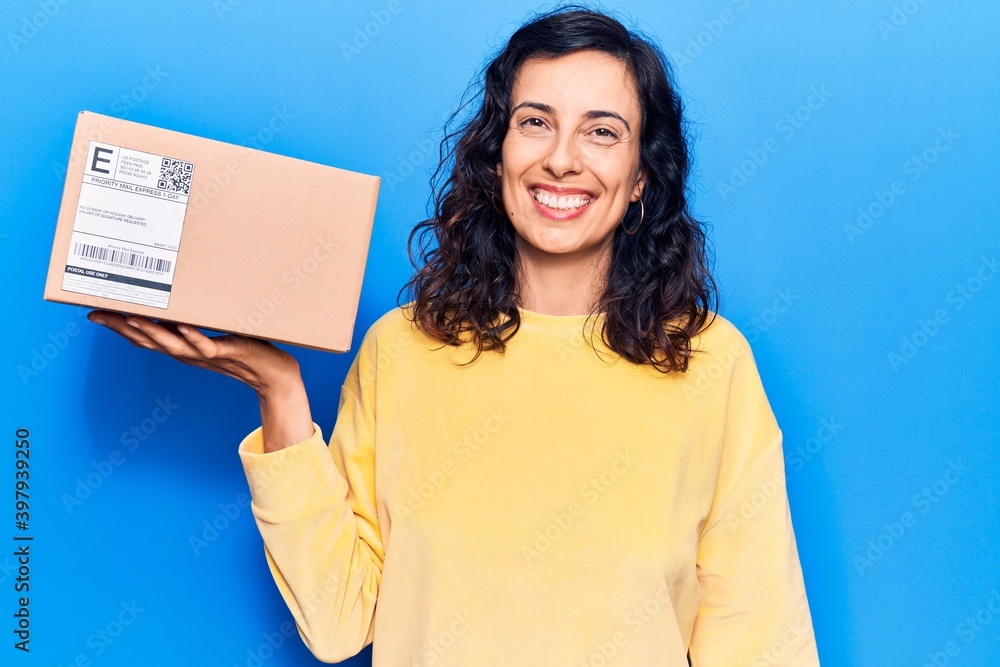 Obraz premium Young beautiful hispanic woman holding delivery package looking positive and happy standing and smiling with a confident smile showing teeth